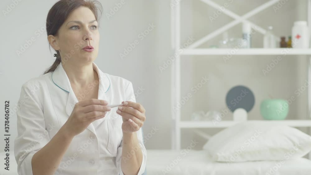 Anxious woman taking temperature and sighing. Portrait of stressed Caucasian doctor having symptoms of Covid-19 viral infection. Risk of medical employees during coronavirus pandemic outbreak. Anxious woman taking temperature and sighing. Portrait of stressed Caucasian doctor having symptoms of Covid-19 viral infection. Risk of medical employees during coronavirus pandemic outbreak.