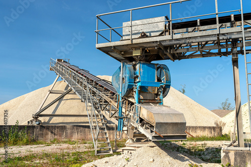 A system of interconnected conveyor belts over heaps of gravel against a blue sky at an industrial cement plant.