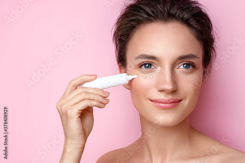 Beautiful woman using cream around the eyes. Photo of woman with perfect makeup on pink background. Beauty and skin care concept