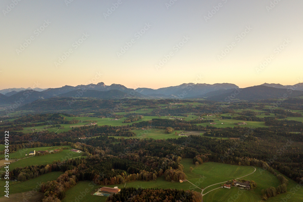 Naklejka premium Aerial view of small scattered farm houses with red tiled roofs among green farming fields and distant mountain forest in summer.