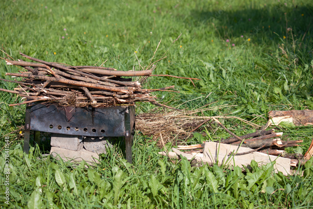 Fototapeta premium Metal grill with firewood among the green grass in the meadow. Near firewood and grass for lighting.