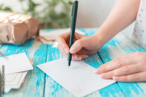 A woman writes on an empty postcard. Hands holding a pen close-up. Blue wooden table with a parcel in the background. The concept of mail correspondence and postcrossing photo