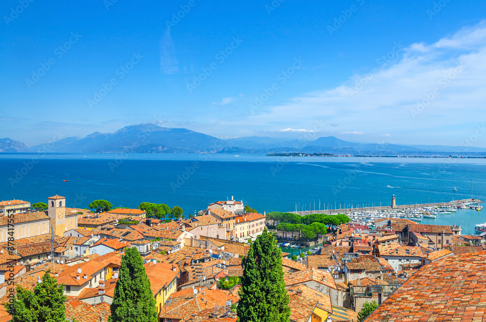 Aerial panoramic view of Desenzano del Garda town, red tiled roof buildings, Garda Lake water surface, Monte Baldo mountain range, Sirmione peninsula, molo pier lighthouse, Lombardy, Northern Italy