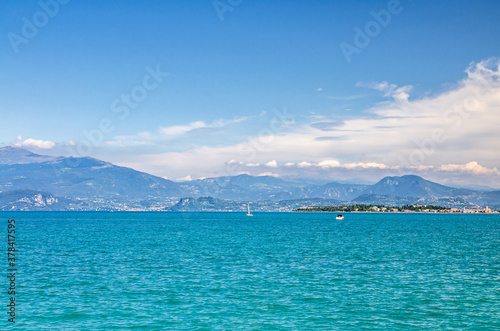 Garda Lake azure turquoise water surface with view of Monte Baldo mountain range and Sirmione peninsula, blue sky white clouds background, Desenzano del Garda town, Lombardy, Northern Italy