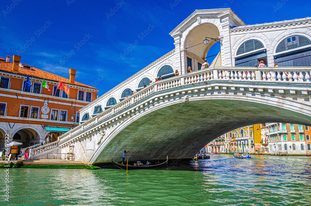 Venice cityscape with Rialto Bridge across Grand Canal waterway, Venetian architecture colorful buildings, gondolas boats sailing Canal Grande, blue sky in sunny day. Veneto Region, Northern Italy.