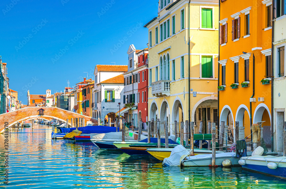 Chioggia cityscape with narrow water canal Vena with moored multicolored boats between old colorful buildings and brick bridge, blue sky background in summer day, Veneto Region, Northern Italy