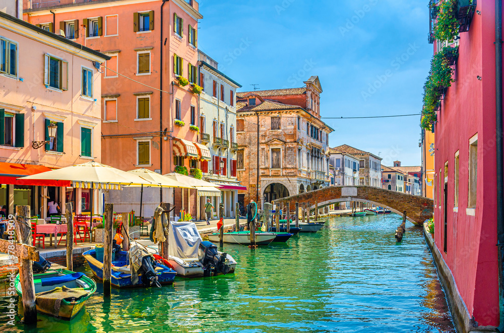 Chioggia cityscape with narrow water canal Vena, moored multicolored boats, street restaurant on embankment, old colorful buildings and brick bridge, blue sky in summer day, Veneto Region, Italy