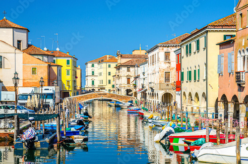 Chioggia cityscape with narrow water canal Vena with moored multicolored boats between old colorful buildings and brick bridge, blue sky background in summer day, Veneto Region, Northern Italy © Aliaksandr