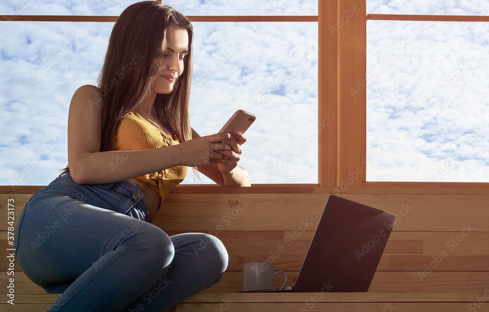 Woman using smartphone and laptop while sitting on window sill Woman using smartphone and laptop while sitting on window sill