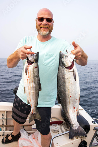 Happy bald man wearing sunglasses holds two fresh caught Coho salmon fish on the Puget Sound in Washington State