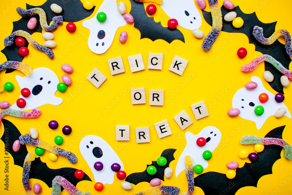 inscription from wooden blocks trick or treat and frame made of paper homemade bats and paper ghosts and multicolored candies and worms from gummy