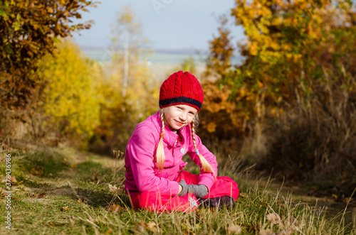 adorable blond girl with red cap in autumnal nature in colorful sunny day
