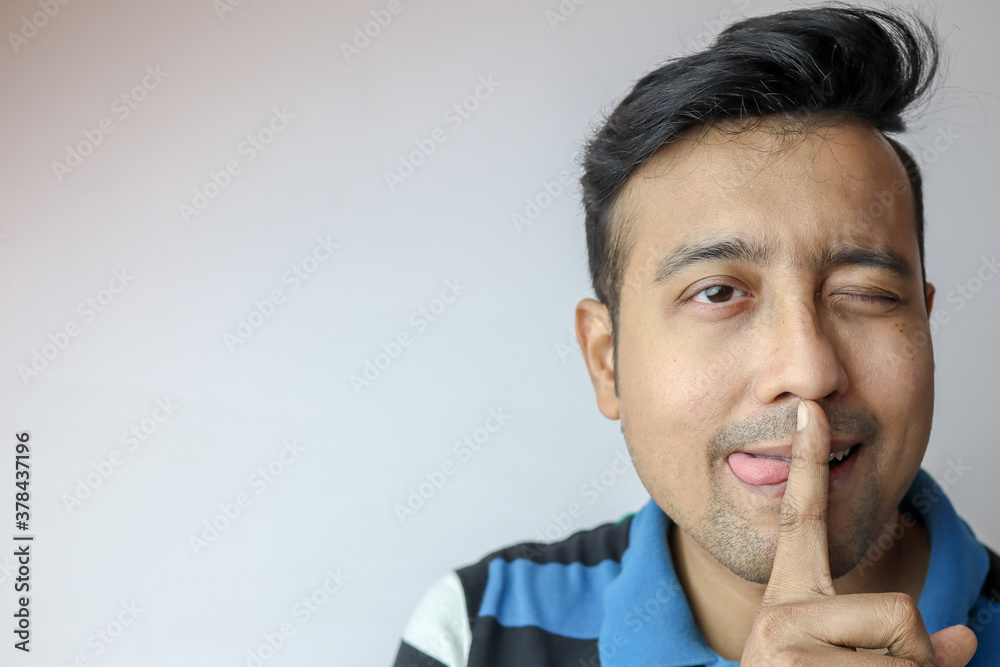 a young male winking with tongue out of mouth and gesturing to keep quiet and a funny expression on face isolated on grey with copy space for text. a young male winking with tongue out of mouth and gesturing to keep quiet and a funny expression on face isolated on grey with copy space for text.