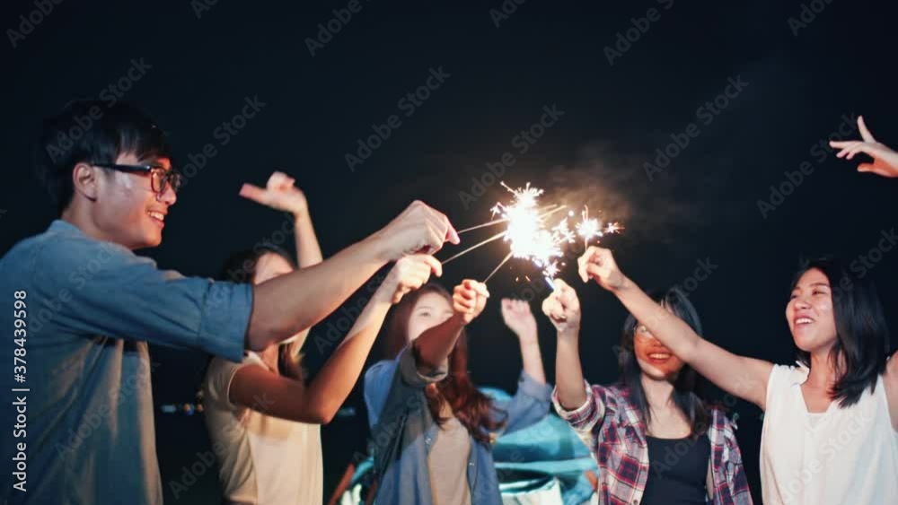Group of young Asian college student friends lit light sparkler, sing and dance together at beach camping tent. Party people, love friendship relationship, or outdoor camping activity concept