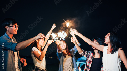 Group of young Asian college student friends lit light sparkler, sing and dance together at beach camping tent. Party people, love friendship relationship, or outdoor camping activity concept