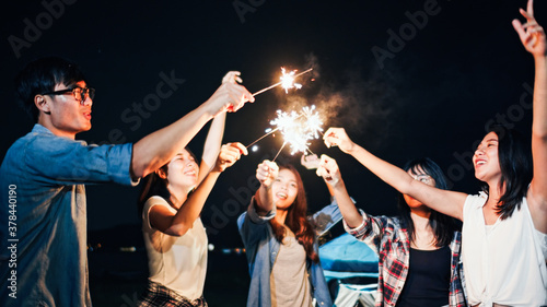 Group of young Asian college student friends lit light sparkler, sing and dance together at beach camping tent. Party people, love friendship relationship, or outdoor camping activity concept