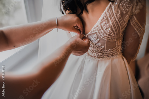 The back of the bride in a wedding dress. Mom's and woman's hands help fasten the clasps at the back. 