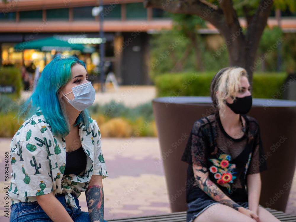 Mujeres jóvenes modernas sentadas en un banco hablando mientras respetando las normas de seguridad y distanciamiento social de la pandemia del coronavirus