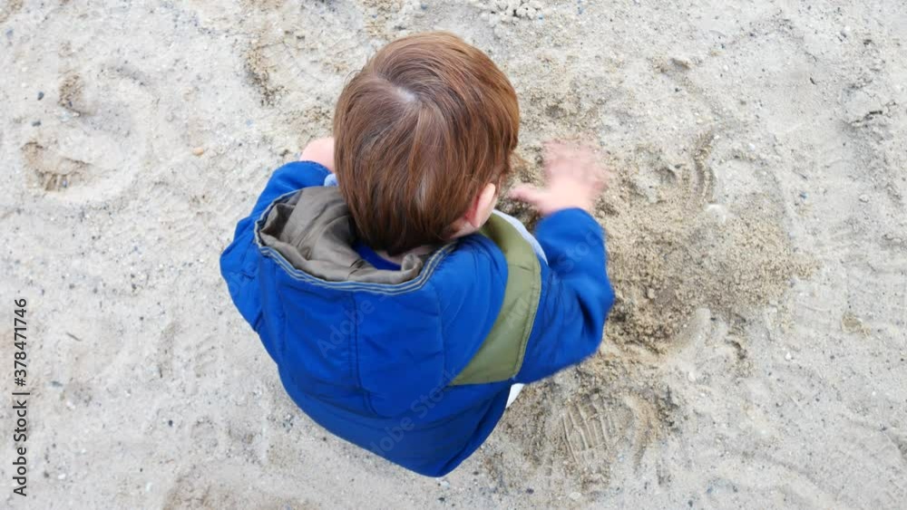 One little boy wearing jacket plays with the sand squatting and then raises his head and looks up One little boy wearing jacket plays with the sand squatting and then raises his head and looks up