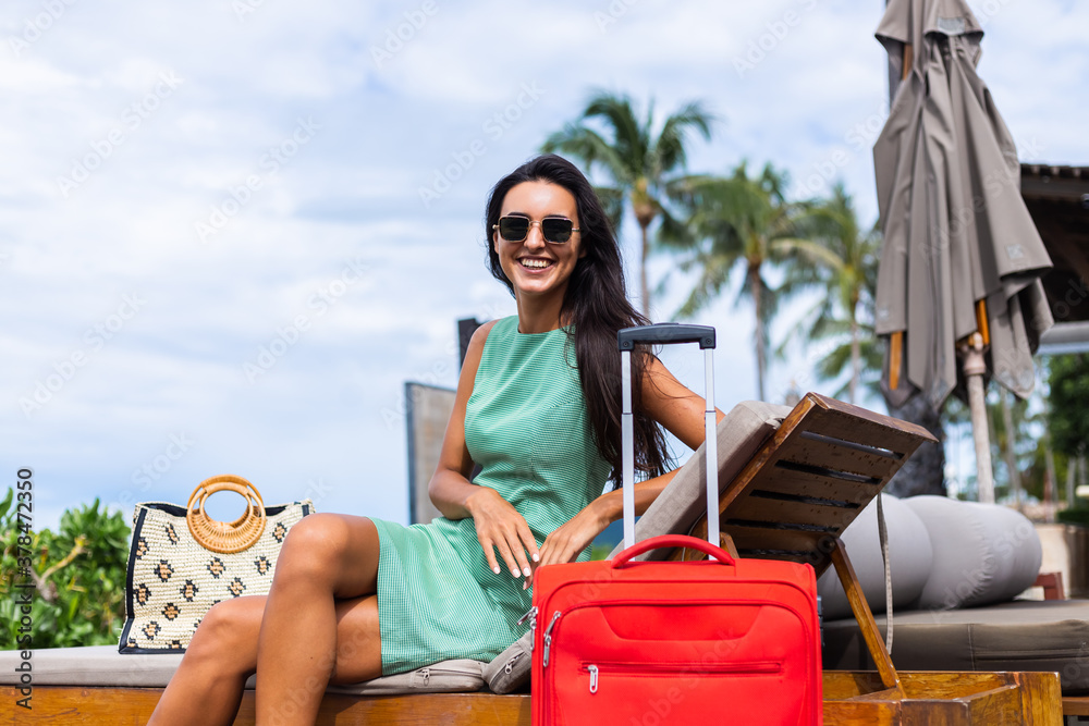 Happy caucasian pretty long hair elegant tourist woman in dress with red suitcase outside hotel on vacation. Travel concept.  