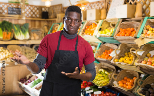 Friendly African American seller meeting customers in greengrocery store, offering fresh fruits and vegetables for sale