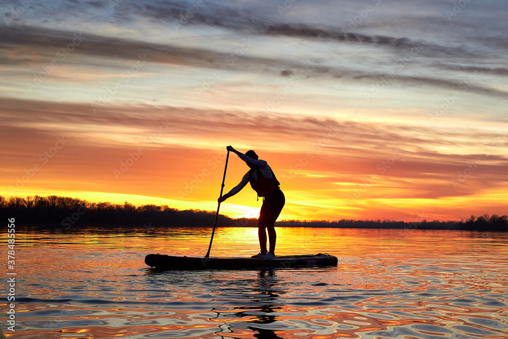 Silhouette of woman paddle on stand up paddle boarding (SUP) on quiet winter or autumn river at sunset. Colorful sunset over the river