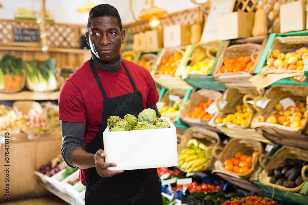 Confident african american salesman carrying box with fresh artichokes at vegetable market