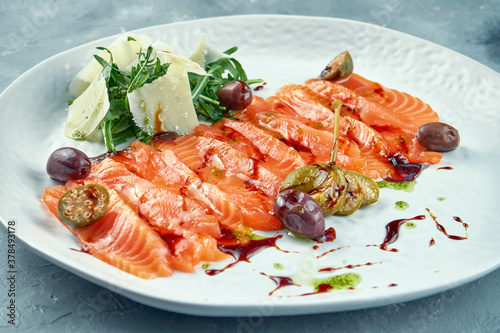 Sliced lightly salted salmon with arugula and parmesan. Red fish sashimi with capers and olives on a white plate on a gray background.. Fresh seafood