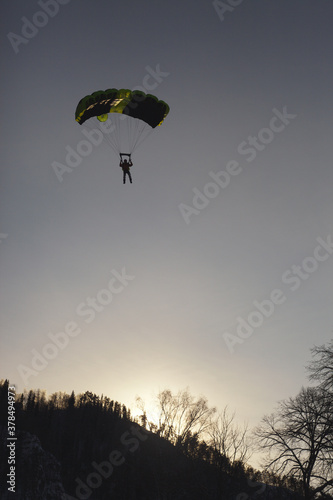 Silhouette of a BASE jumper under a parachute canopy against the background of a hillside in the evening light.