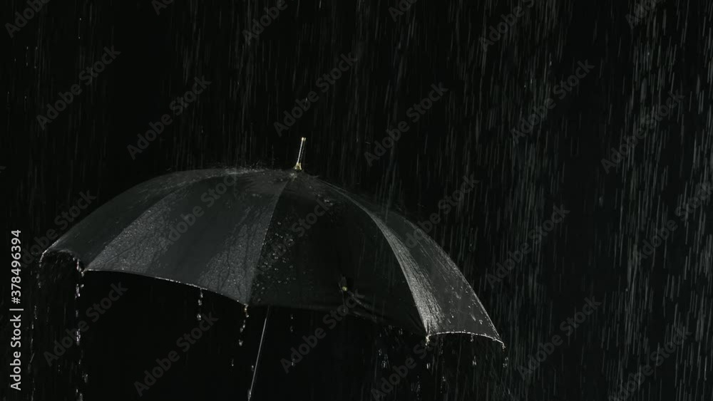 An open black umbrella in the rain through which streams of water flow down. Filmed close up in a dark backlit studio. Slow motion.