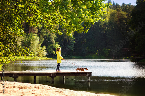 A girl walks her dog in the Park. A child and a red Dachshund on a leash stands on a bridge near the water in autumn in Sunny weather.