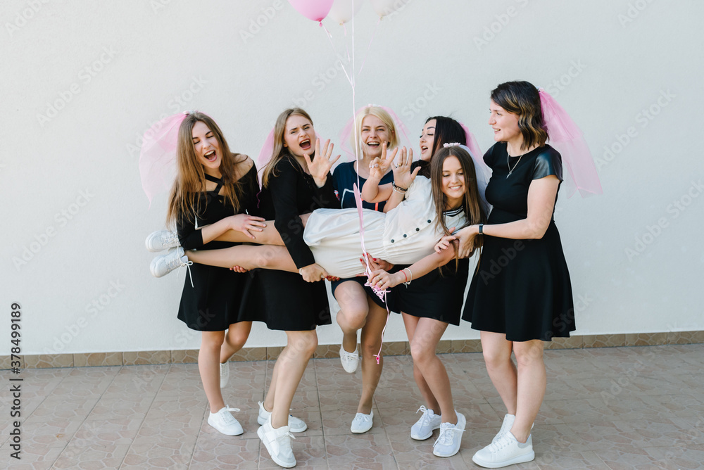 Fototapeta premium Bride and bridesmaids stands near white wall outdoors and celebrating bachelorette party. Beautiful sexy young girls in dress and veil on bachelor party. Maiden evening, Hen-parties. Place for text.