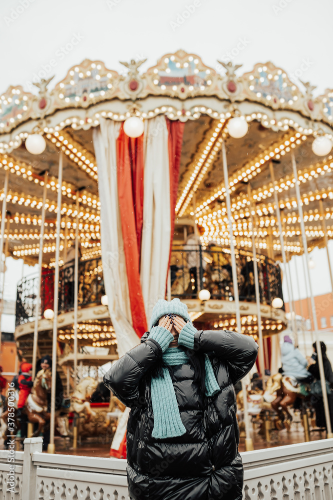 Beautiful girl in stylish fashion jacket and knitted hat and scarf happy covers his face with his hands near the carousel in amusement park.