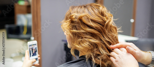Hairdresser doing hairstyle for young woman with red curly hair and with smartphone in her hands in beauty salon