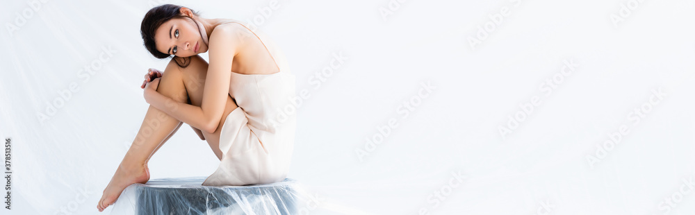 panoramic crop of barefoot woman in silk dress sitting on stool wrapped in plastic bag on white, ecology concept panoramic crop of barefoot woman in silk dress sitting on stool wrapped in plastic bag on white, ecology concept