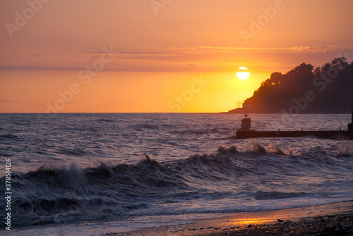 Stormy sea waves on the seashore against the pear and backdrop of the setting sun and sky with clouds. © Alexander