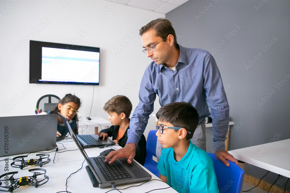 Pupils doing task on laptops and focused teacher monitoring them. Cute children sitting at table, using computers during lesson and studying at school. Knowledge and digital education concept Pupils doing task on laptops and focused teacher monitoring them. Cute children sitting at table, using computers during lesson and studying at school. Knowledge and digital education concept