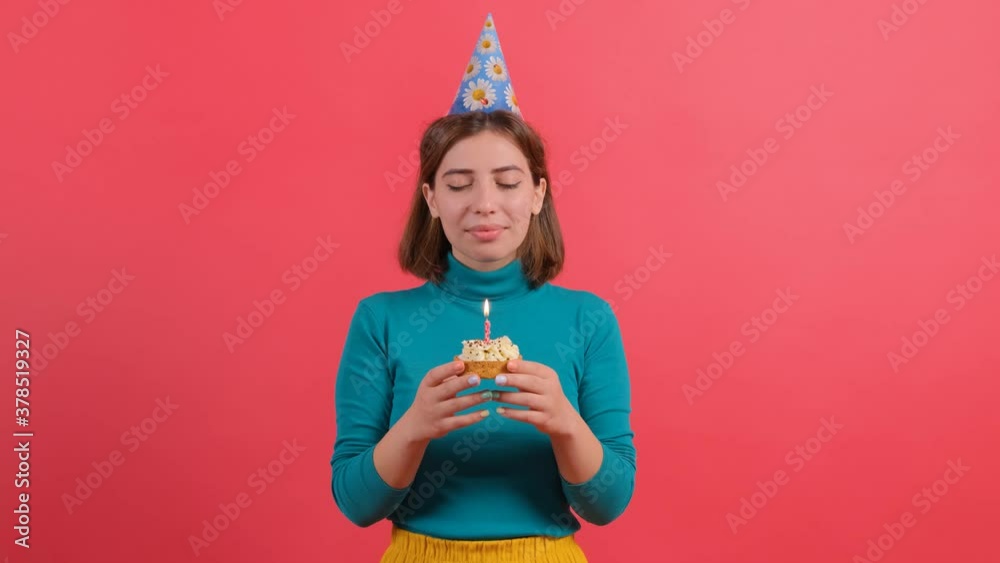 Side view of young woman in knitted pink sweater, birthday hat blowing out candle on cake in hands isolated on blue turquoise background studio portrait. People lifestyle concept. Mock up copy space Side view of young woman in knitted pink sweater, birthday hat blowing out candle on cake in hands isolated on blue turquoise background studio portrait. People lifestyle concept. Mock up copy space