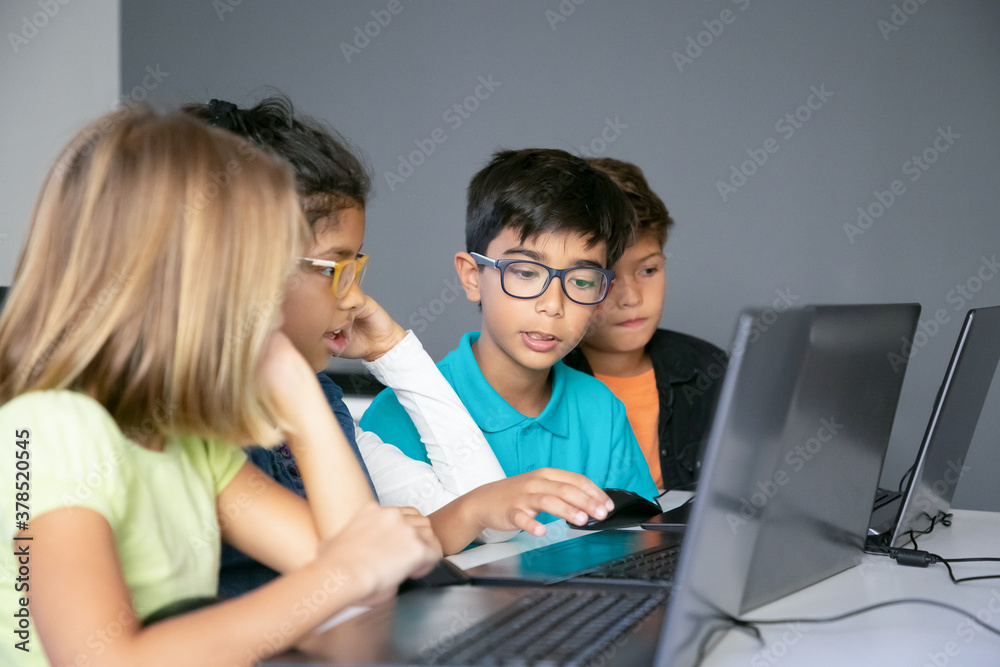Little classmates discussing lesson and doing task. Mixed-race boy looking at laptop screen and talking with school friends about study. Childhood, communication and digital education concept Little classmates discussing lesson and doing task. Mixed-race boy looking at laptop screen and talking with school friends about study. Childhood, communication and digital education concept