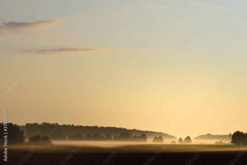 In a wide landscape in Bavaria at dawn the fog pulls up over fields and trees.