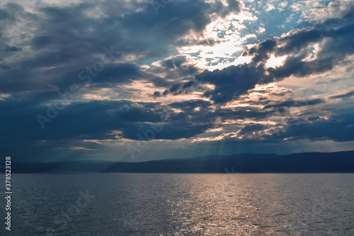 evening bright yellow pink sunset with silvery gray blue clouds on lake baikal with a mountains ridge on the horizon, sun rays