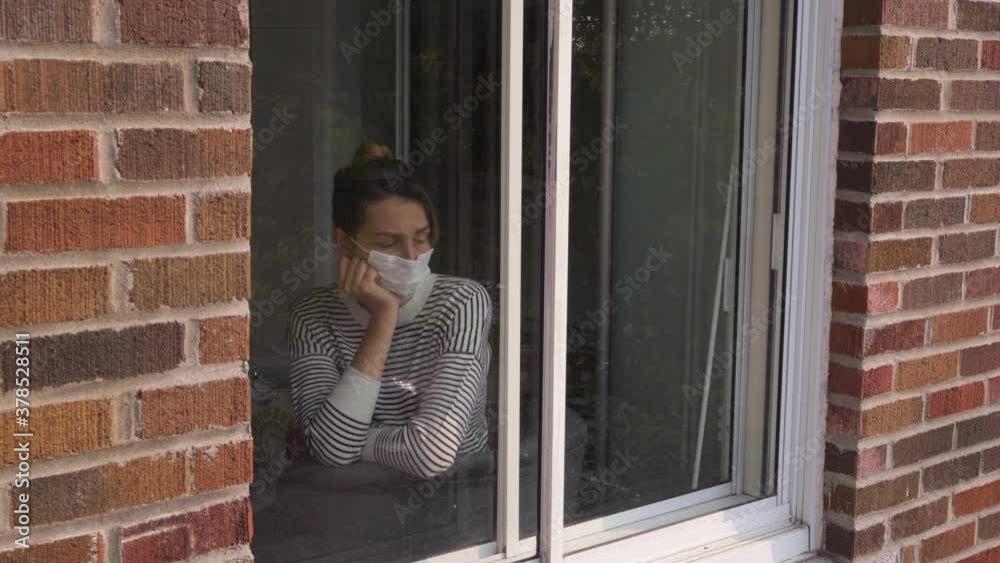Young Woman in medical mask looking through window. Sad and bored girl looks outside on self isolation in quarantine 4K. Stay at home due to Coronavirus Outbreak. Covid 19. Young Woman in medical mask looking through window. Sad and bored girl looks outside on self isolation in quarantine 4K. Stay at home due to Coronavirus Outbreak. Covid 19.