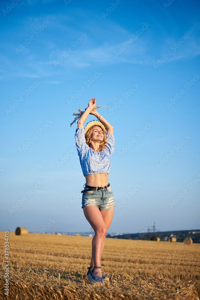 Young curly blond woman, wearing jeans shorts and light blue shirt, holding straw hat, standing on bale on field in summer. Female portrait in natural rural scene. Environmental eco tourism concept. Young curly blond woman, wearing jeans shorts and light blue shirt, holding straw hat, standing on bale on field in summer. Female portrait in natural rural scene. Environmental eco tourism concept.