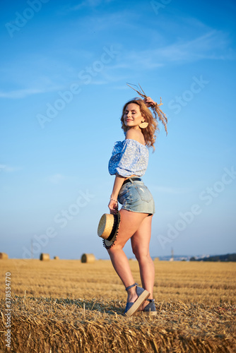 Young curly blond woman, wearing jeans shorts and light blue shirt, holding straw hat, standing on bale on field in summer. Female portrait in natural rural scene. Environmental eco tourism concept.