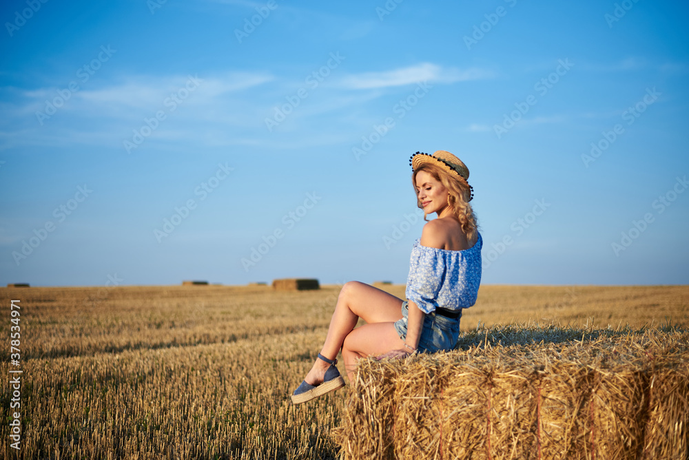 Young curly blond woman, wearing jeans shorts, light blue shirt and straw hat, sitting on bale on field in summer. Female portrait in natural rural scene. Environmental eco tourism concept. Young curly blond woman, wearing jeans shorts, light blue shirt and straw hat, sitting on bale on field in summer. Female portrait in natural rural scene. Environmental eco tourism concept.