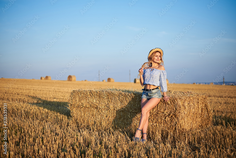 Young curly blond woman, wearing jeans shorts, light blue shirt and straw hat, sitting on bale on field in summer. Female portrait in natural rural scene. Environmental eco tourism concept. Young curly blond woman, wearing jeans shorts, light blue shirt and straw hat, sitting on bale on field in summer. Female portrait in natural rural scene. Environmental eco tourism concept.