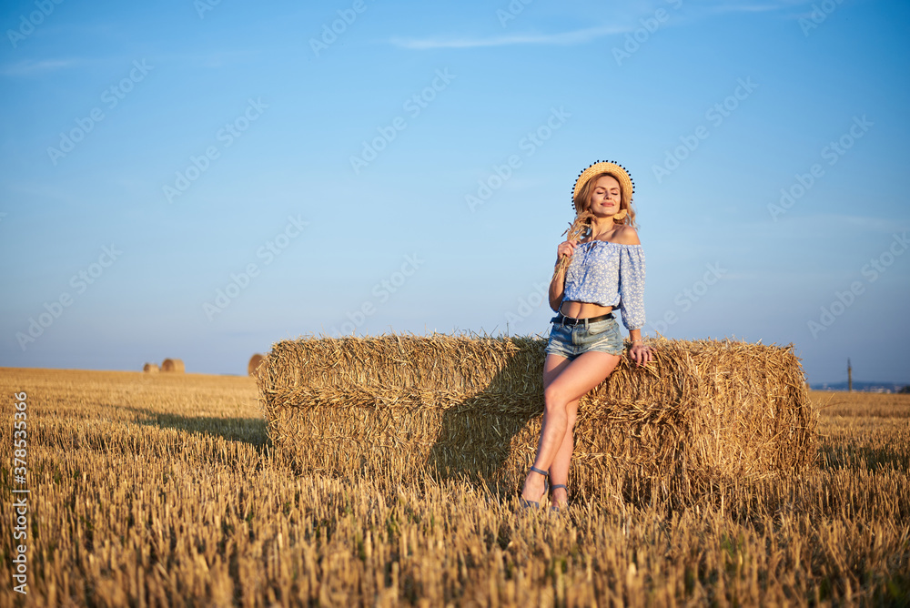 Young curly blond woman, wearing jeans shorts, light blue shirt and straw hat, sitting on bale on field in summer. Female portrait in natural rural scene. Environmental eco tourism concept. Young curly blond woman, wearing jeans shorts, light blue shirt and straw hat, sitting on bale on field in summer. Female portrait in natural rural scene. Environmental eco tourism concept.