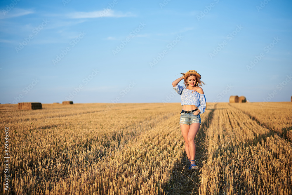 Young curly blond woman, wearing jeans shorts, light blue shirt and straw hat, walking running jumping on field in summer. Female portrait in natural rural scene. Environmental eco tourism concept. Young curly blond woman, wearing jeans shorts, light blue shirt and straw hat, walking running jumping on field in summer. Female portrait in natural rural scene. Environmental eco tourism concept.