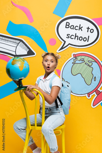 Excited schoolkid holding globe on chair near paper elements on yellow background