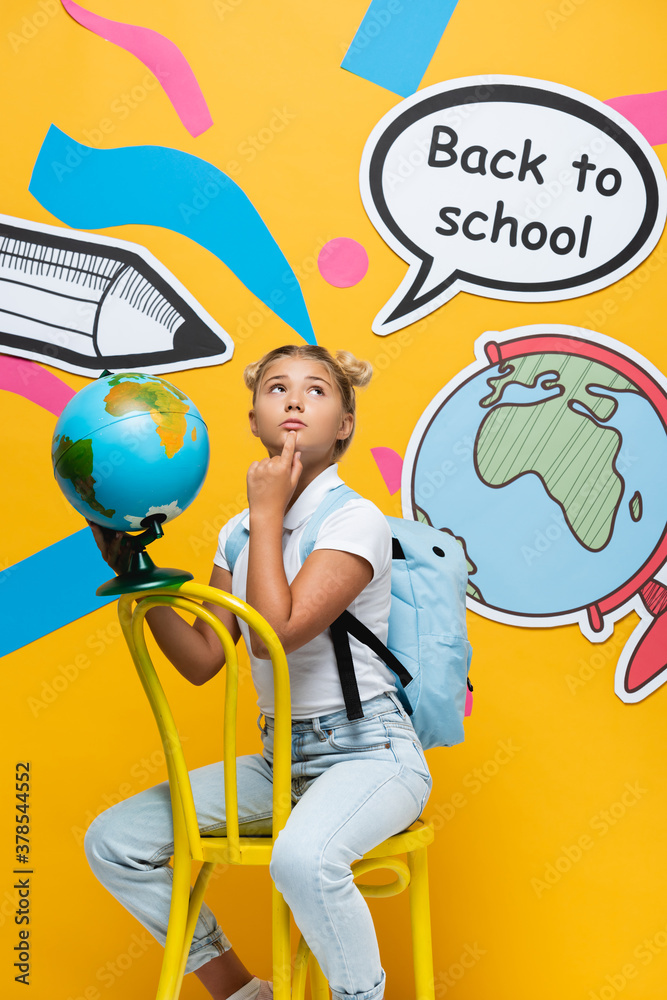 Fototapeta premium Pensive child holding globe near speech bubble with back to school lettering and paper art on yellow background